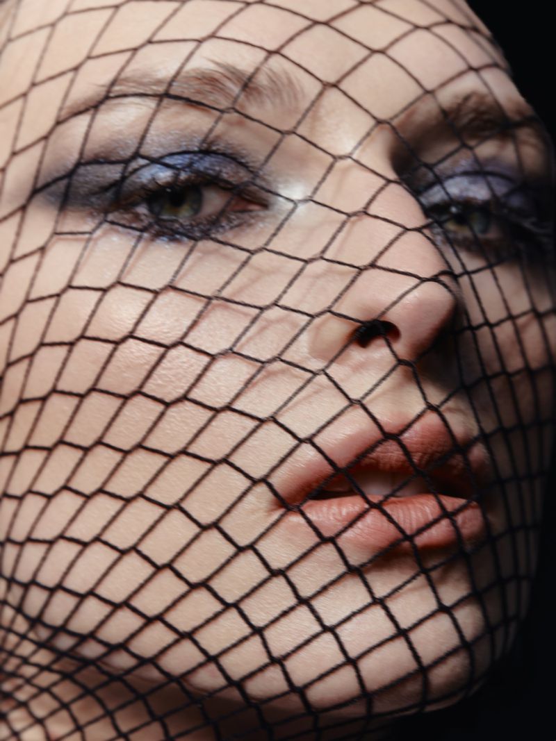 Close-up portrait of a woman's face covered by a black fishnet, featuring soft lighting, and focus on the eyes with blue eyeshadow.