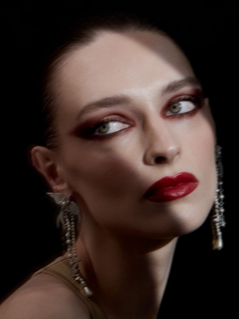 Close-up beauty shot of a young adult woman with dramatic eye makeup, red lipstick, and ornate dangling earrings against a black background.