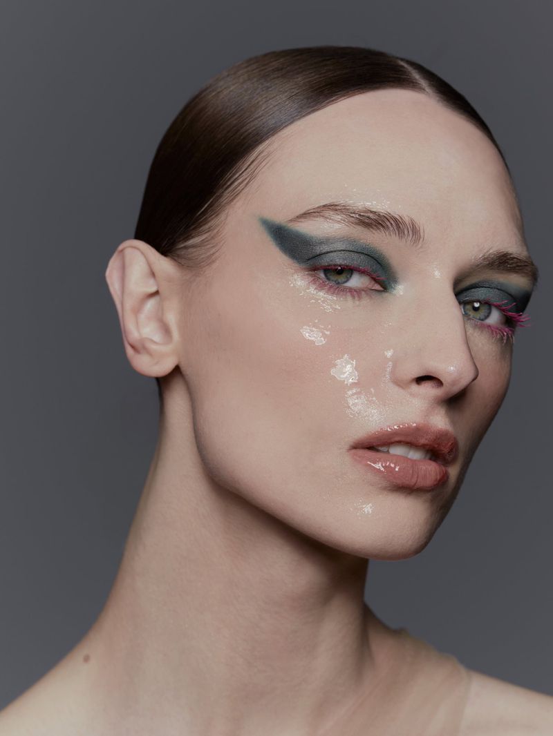 Close-up of a woman with slicked-back hair and dramatic teal and pink eye makeup, posing against a grey backdrop in a beauty shot.