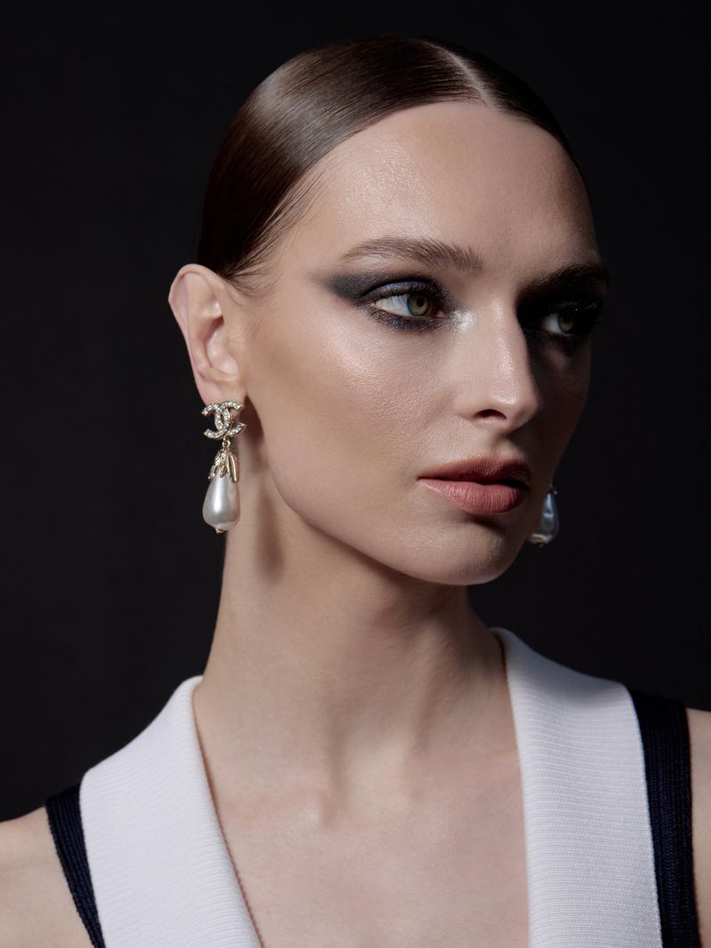 Close-up portrait of a woman with dark eye makeup and slicked-back dark hair wearing designer earrings against a black background.