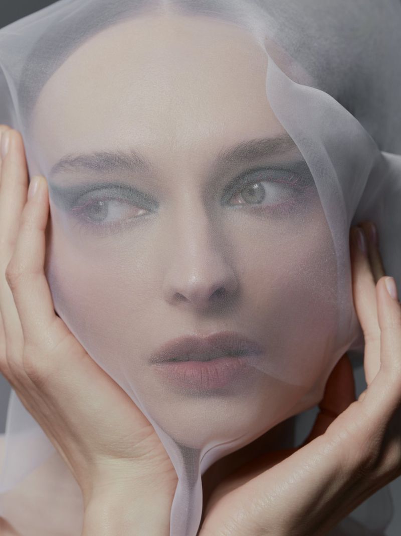 Close-up portrait of a woman with fair skin, light eyes, and dark eye makeup, wearing a sheer white veil and hands near her face.