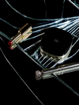 Still life shot of makeup and cosmetic products including lipstick, compact powder, and eyeliner on reflective, cracked glass surface.