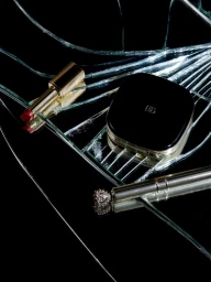 Gold lipstick, black compact case, and metallic cosmetic tool arranged across shattered glass over a black background.