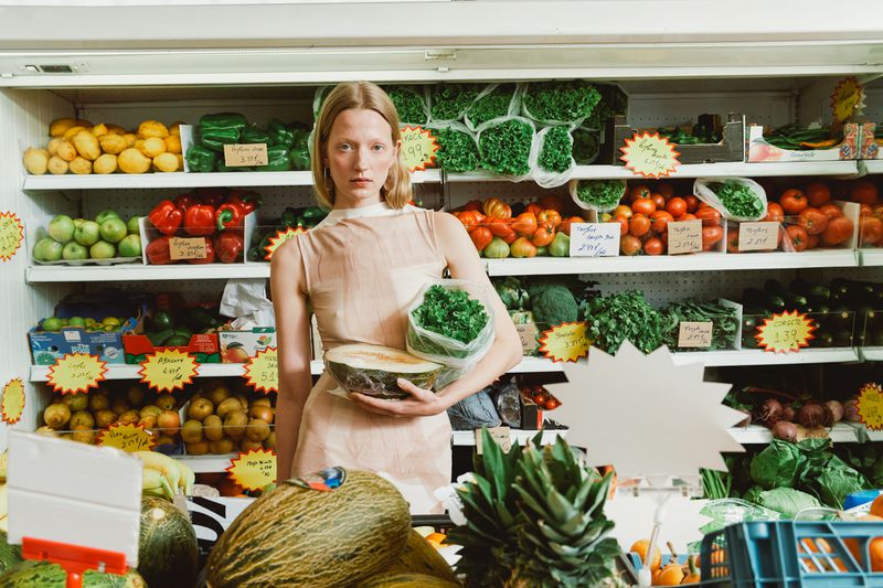 Woman in grocery store holds cantaloupe and bag of greens; shelves with produce like peppers, lettuce, tomatoes in background.