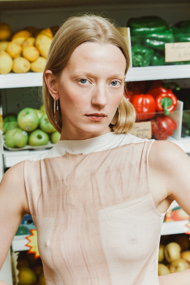 Young woman with fair skin and blonde hair poses in front of a produce display in a grocery store, wearing a transparent top.