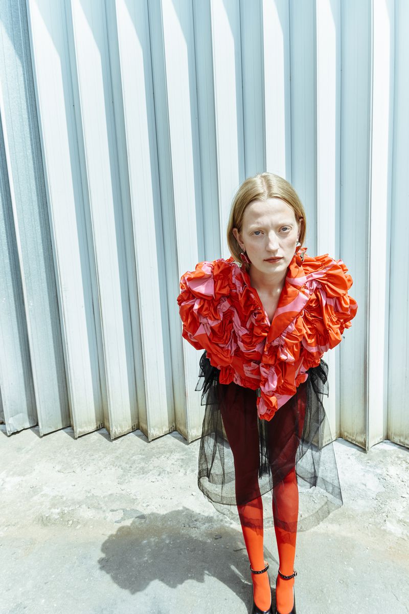 Woman poses in a ruffled orange top and black tulle skirt against a corrugated wall. She's wearing red tights and black heels in a high-angle shot.