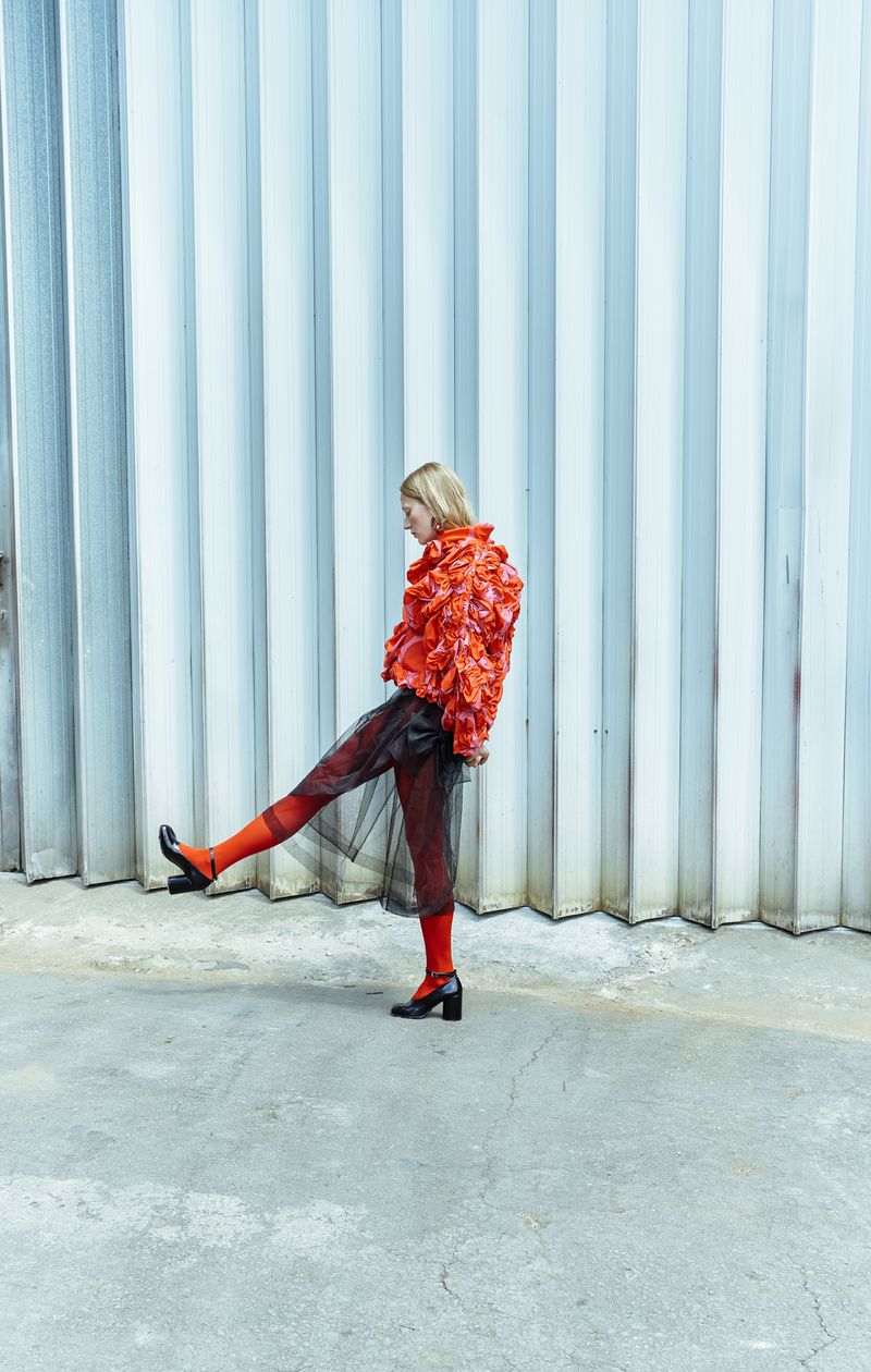 A fashionable woman in an orange floral jacket, black sheer skirt, and orange stockings strikes a pose against a textured metal wall.