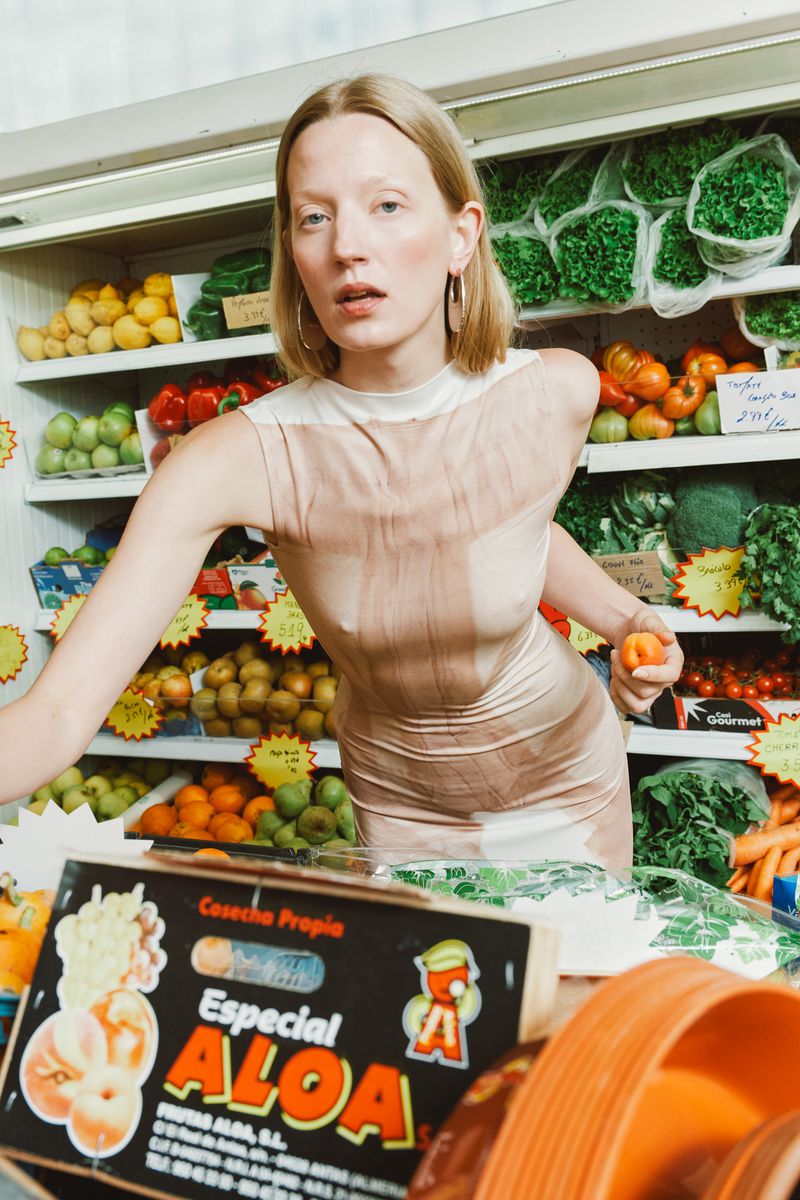 A blonde woman in a dress stands in a produce aisle holding an orange, with a variety of fresh vegetables and fruits in the background.