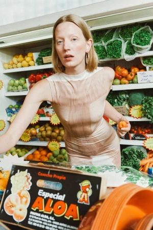 A blonde woman in a dress stands in a produce aisle holding an orange, with a variety of fresh vegetables and fruits in the background.