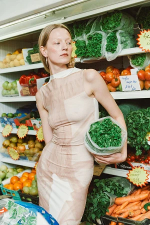 Blonde woman in a tan dress holding a bag of leafy greens in a grocery store aisle surrounded by fresh produce on shelves.