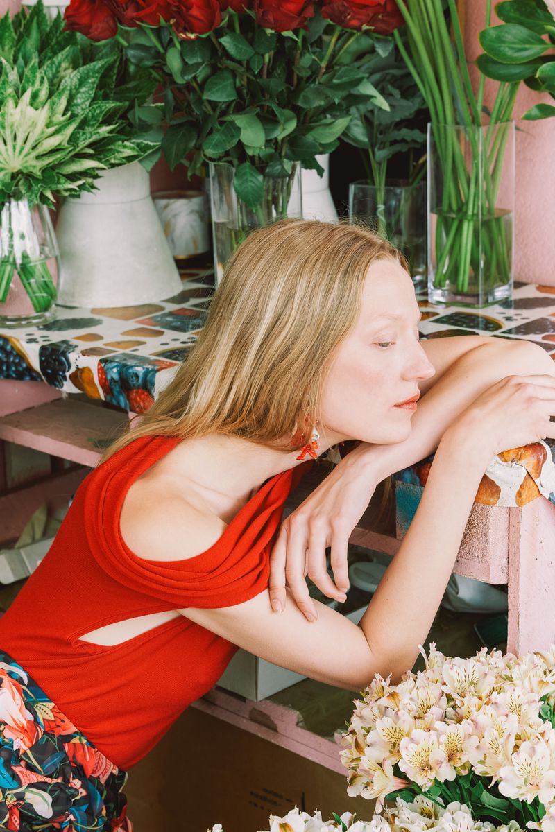 Adult woman in a red top leans against a table surrounded by flowers in vases in an indoor setting.