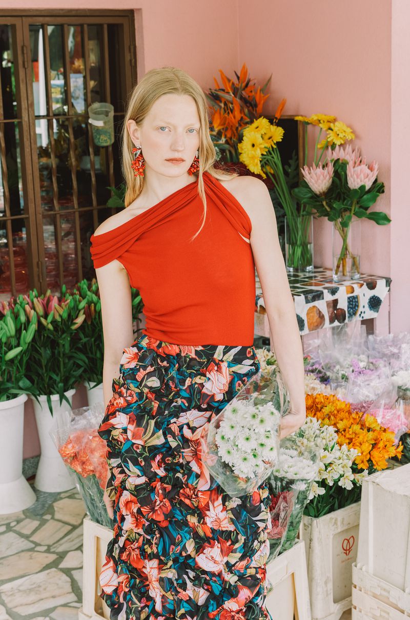 A fashionable adult woman with blond hair stands among flowers at a flower stand, wearing a red top and floral skirt.