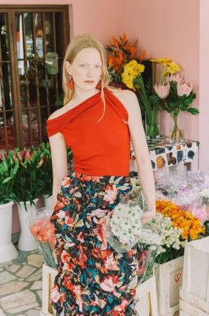 A fashionable adult woman with blond hair stands among flowers at a flower stand, wearing a red top and floral skirt.