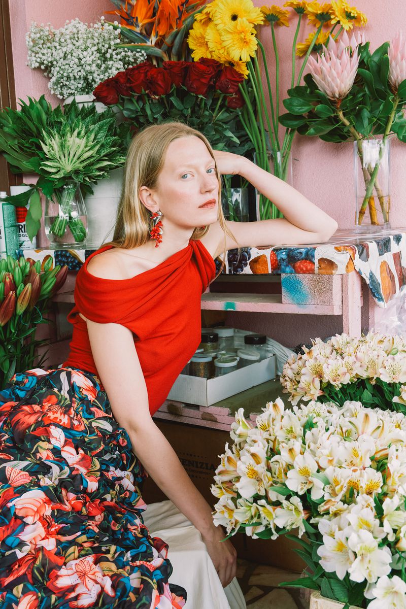 A woman with long blonde hair poses in front of flowers. She's wearing a red top and floral skirt, shot in a brightly lit setting.
