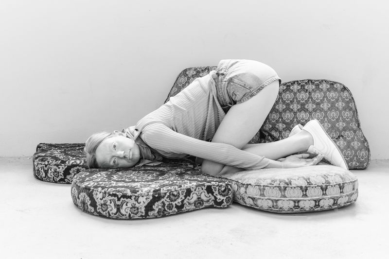 Monochrome image of a fair-skinned woman lying on patterned cushions. She wears jean shorts and a sweater. The setting is a plain indoor studio.