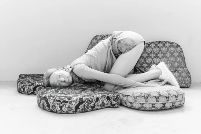Monochrome image of a fair-skinned woman lying on patterned cushions. She wears jean shorts and a sweater. The setting is a plain indoor studio.