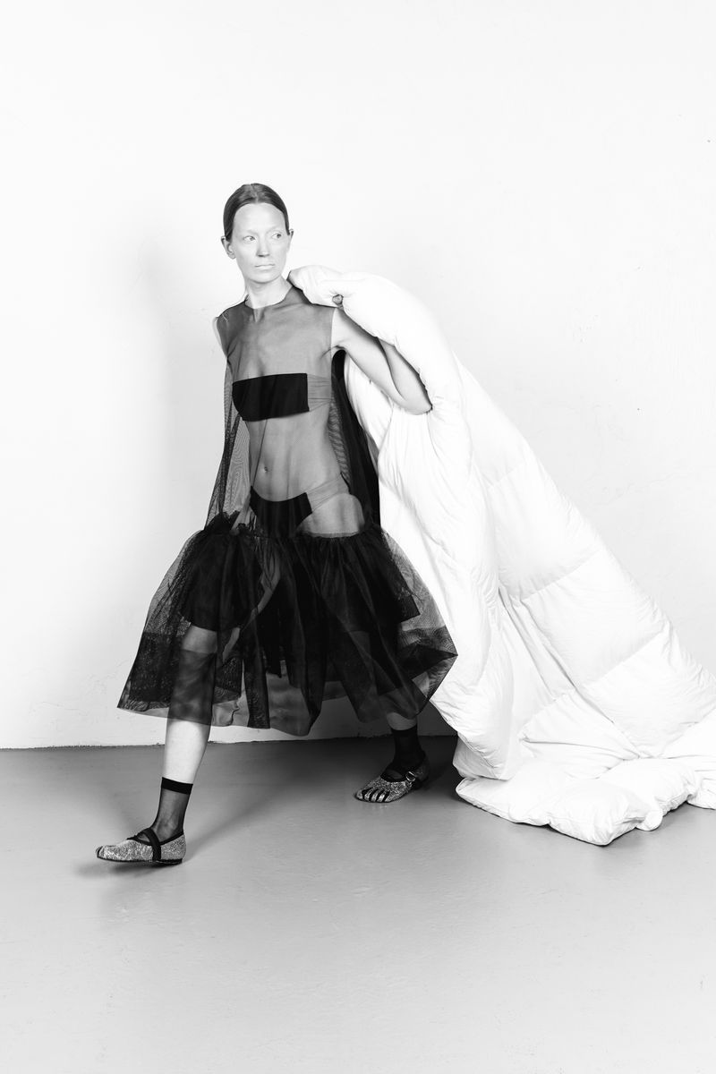 Adult female fashion model posing in sheer black tulle dress with white quilted throw over shoulder in photo studio on grey floor.