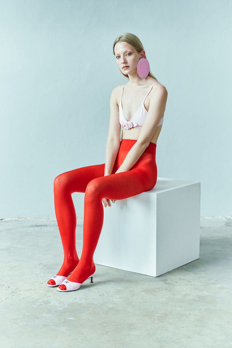Woman in bralette and red tights sitting on a white block, wearing large pink disc earrings and heels against a blue wall.