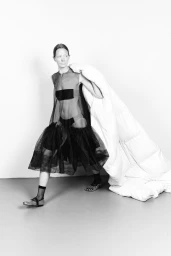 Model in black sheer dress holding a large white puffer coat against a plain backdrop for Sicky Magazine MOVING project