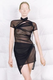 Model Marie Louwes posing in a sheer black draped asymmetric mini dress against a white quilted padded background.