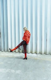 Model wearing a textured red top and dark sheer skirt kicking leg against a corrugated industrial backdrop