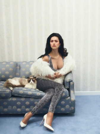 A woman with long dark hair poses on a floral patterned sofa with a cat in a well-lit, carpeted room with striped wallpaper.