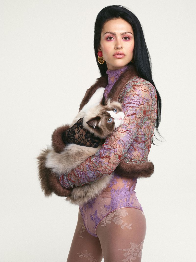 Adult female posing indoors with a long-haired cat. Woman wears a lavender lace bodysuit with long sleeves and fur bolero. White background.