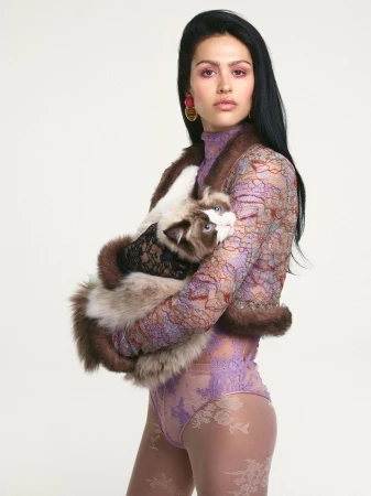Adult female posing indoors with a long-haired cat. Woman wears a lavender lace bodysuit with long sleeves and fur bolero. White background.