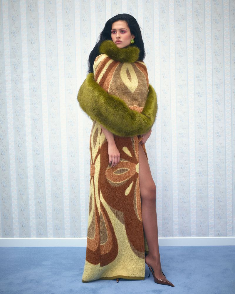 Woman posing in a patterned brown and beige gown with a green faux-fur stole, standing against a vertically striped wallpaper.