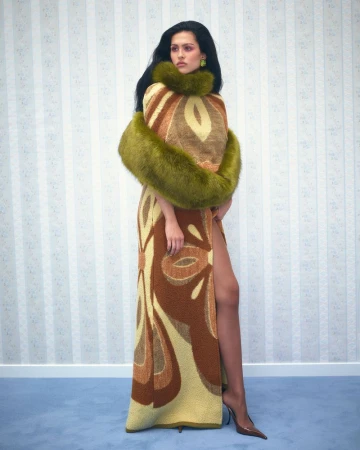 Woman posing in a patterned brown and beige gown with a green faux-fur stole, standing against a vertically striped wallpaper.