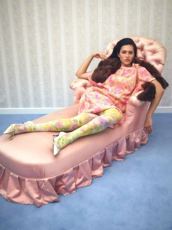 Amelia Gray in a pink floral dress and fur stole reclining on a tufted pink satin chaise lounge over blue carpet.