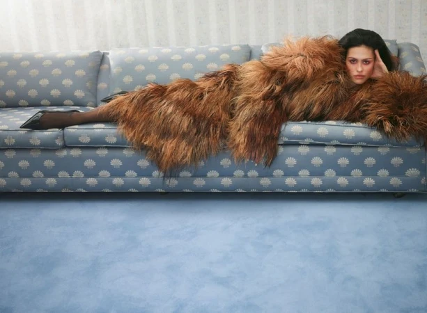 Model Amelia Gray lying across a patterned blue sofa wearing an oversized brown shaggy coat and black pointed high heels.