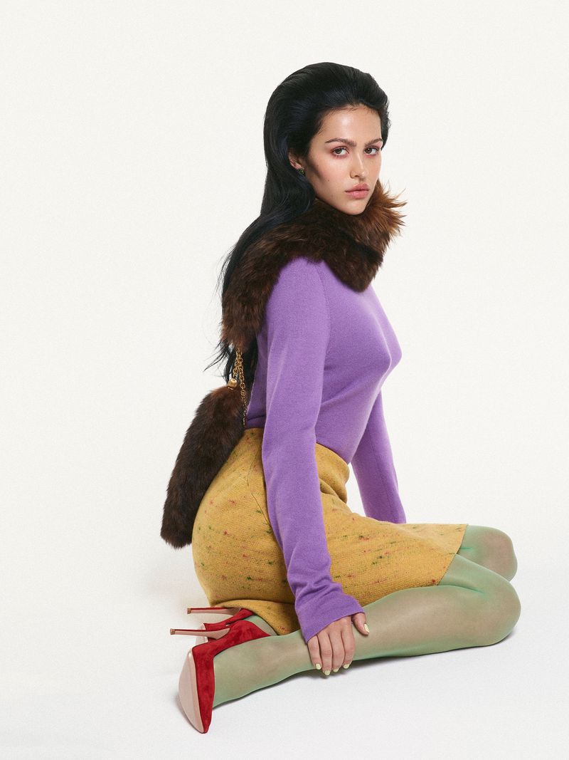 A brunette woman poses in a lavender sweater, mustard skirt, patterned tights, and red heels against a bright white backdrop.
