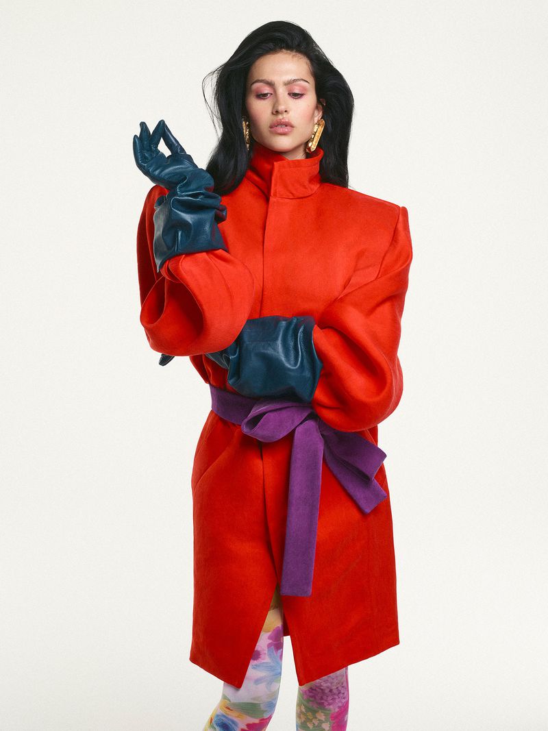 A woman models a vibrant red coat with a purple sash and green gloves against a bright white background in a fashion editorial shot.