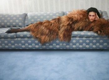 Model Amelia Gray lying across a patterned blue sofa wearing an oversized brown shaggy coat and black pointed high heels.