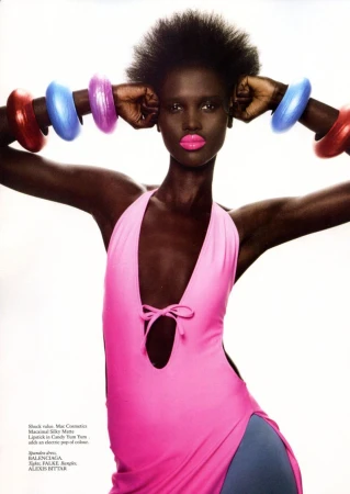 Dark-skinned fashion model wearing pink dress and colorful bangles, posing against a white backdrop in a studio setting.
