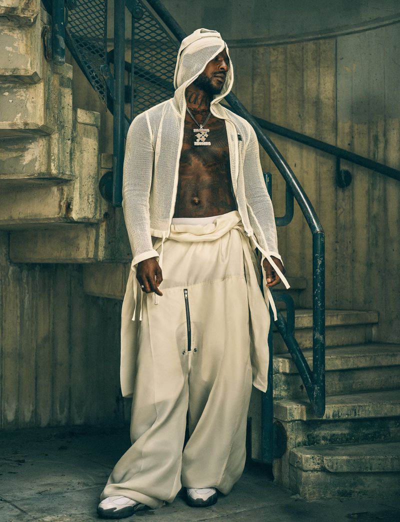 Fashionable man in a hooded sheer shirt and baggy pants poses in front of a weathered staircase with metal railings indoors.