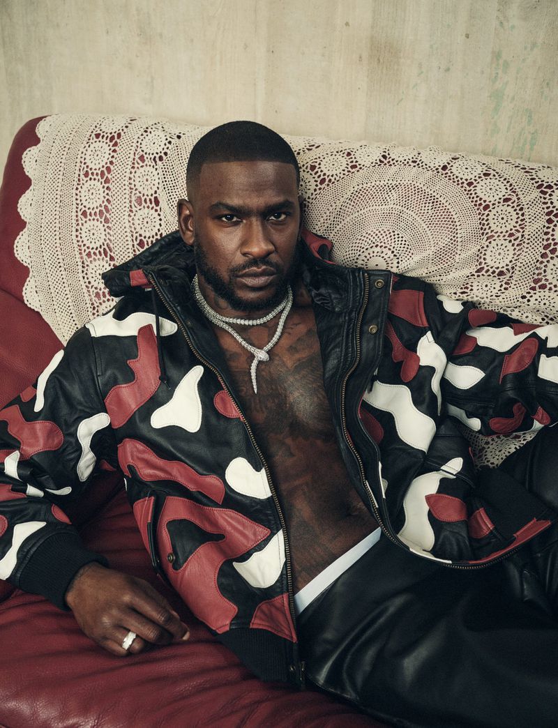 Close-up portrait of a man with short hair, wearing a patterned jacket and jewelry, reclining on a red surface with a patterned backdrop.