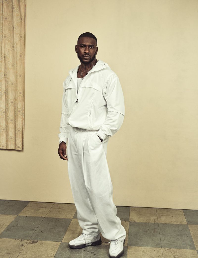 Full-length portrait of an African-American man in a white tracksuit standing indoors with neutral background, relaxed pose, and checkered floor.