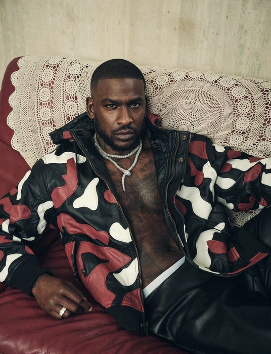 Skepta reclining on a red leather sofa wearing an unzipped camouflage leather jacket and a silver snake chain necklace.