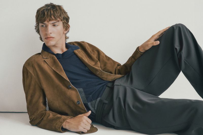 A young adult male model with wavy brown hair reclines on a white surface, wearing a brown jacket, navy shirt, and grey pants.