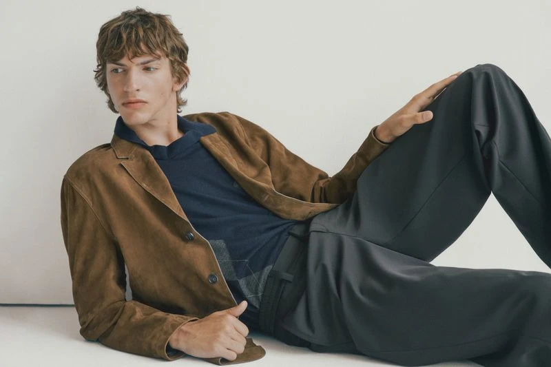 A young adult male model with wavy brown hair reclines on a white surface, wearing a brown jacket, navy shirt, and grey pants.