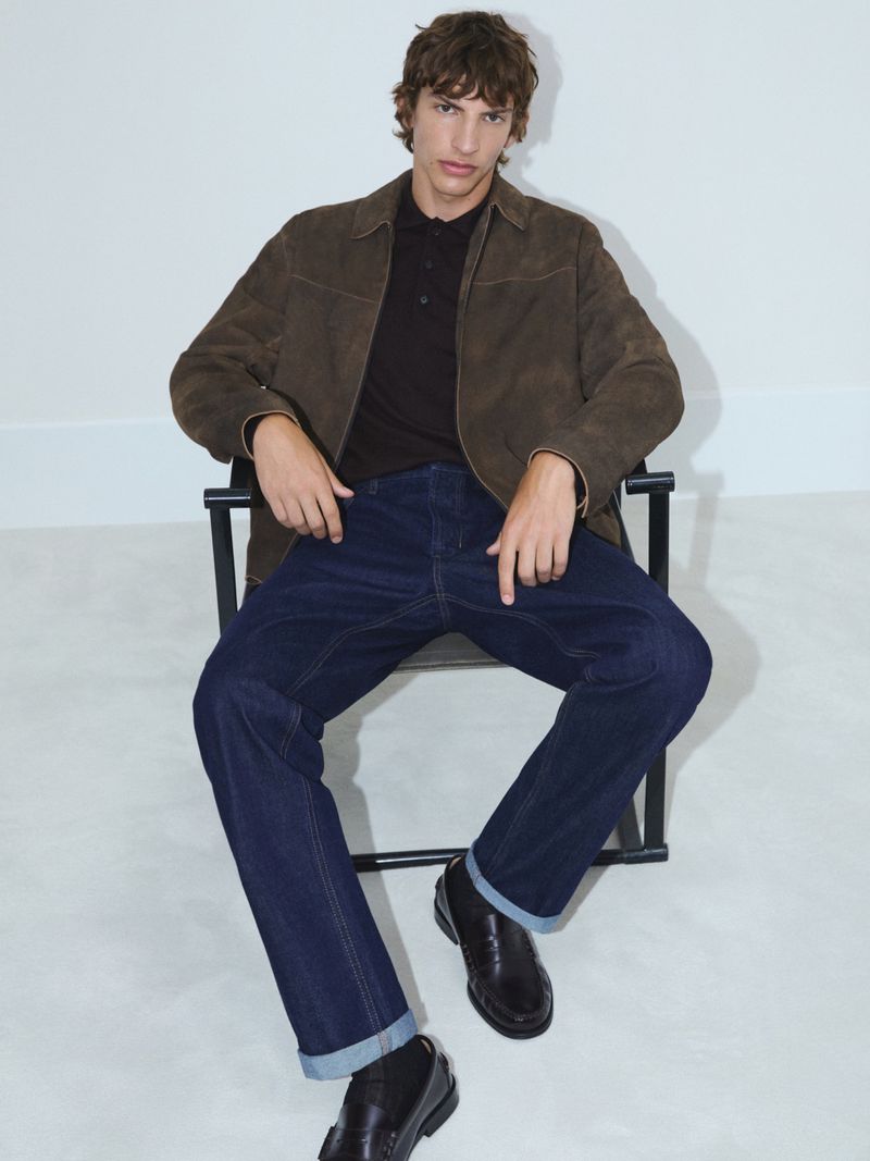 A brunette adult male model poses sitting in a black chair, wearing a brown jacket, dark shirt, blue jeans, and black loafers, in a studio.