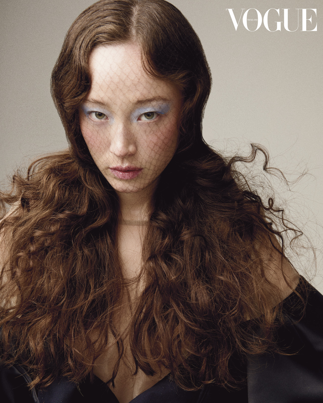 Portrait of a young Asian woman with long wavy brown hair and blue eyeshadow, wearing a black net veil, against a beige background.
