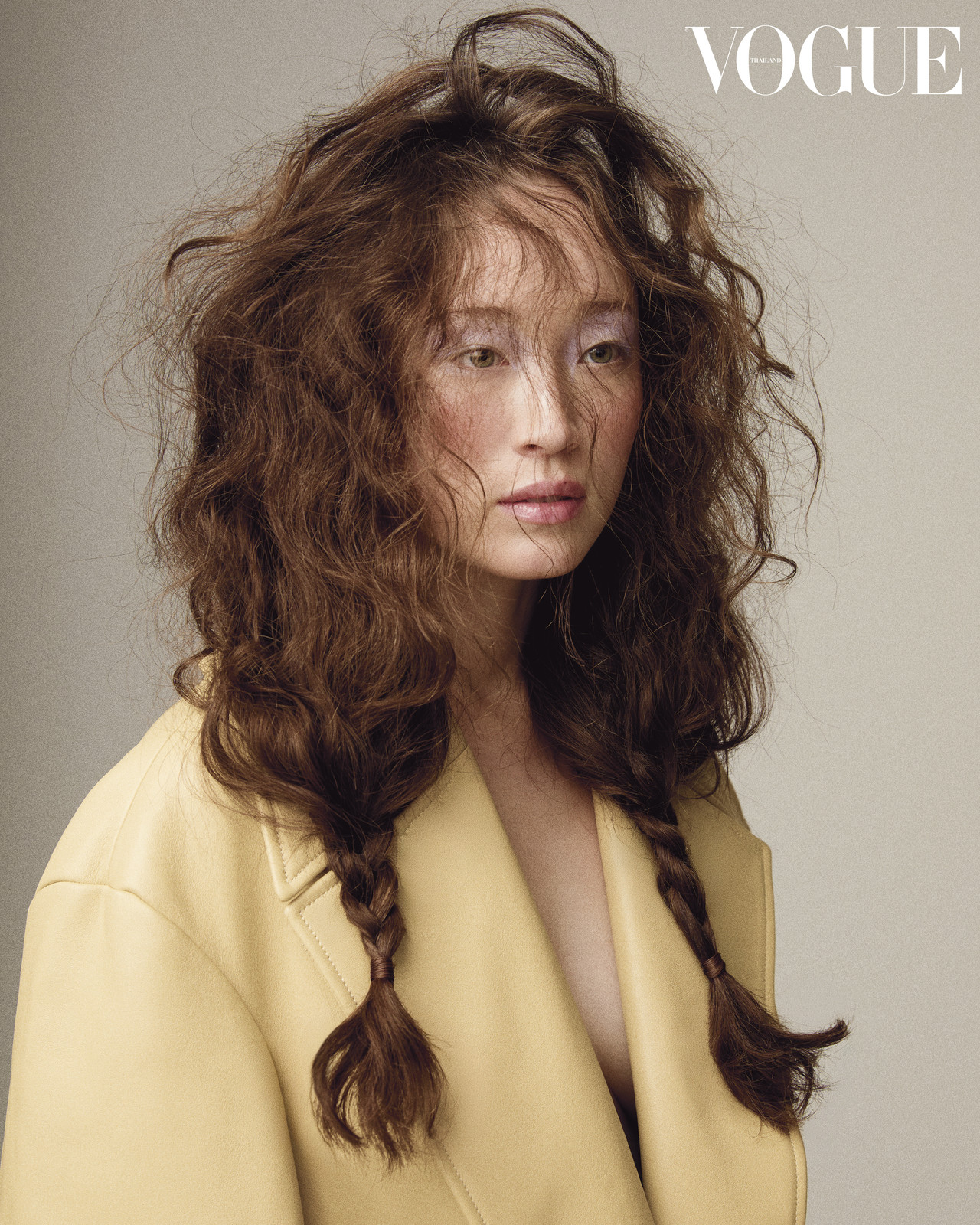 Portrait of a brunette female model with her hair in braids, wearing a yellow coat, and posing against a neutral backdrop for Vogue.