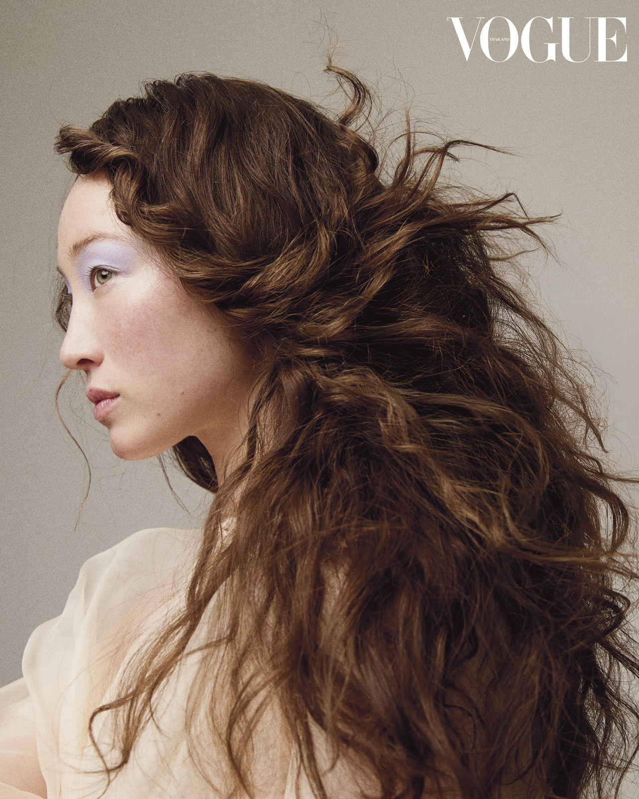 Brunette woman with light purple eyeshadow and styled hair, wearing a neutral-toned garment, as featured in Vogue magazine.