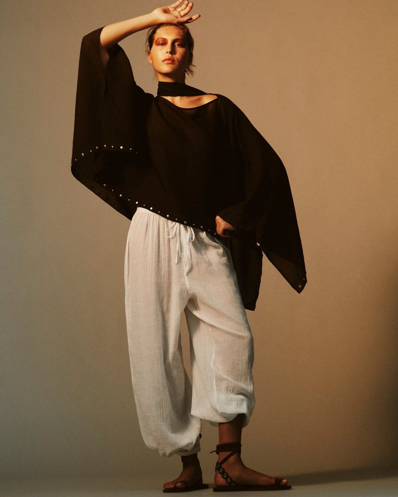 Fashion shot: woman in a black flowing top and white pants poses in a studio with soft, warm lighting and neutral background.