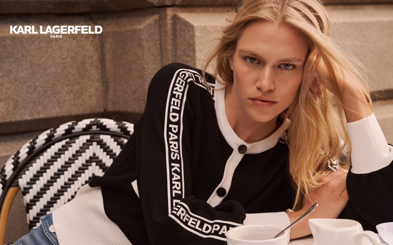 A blonde female model poses at an outdoor cafe wearing a black and white Karl Lagerfeld sweater, sitting at a white table with white coffee cups.