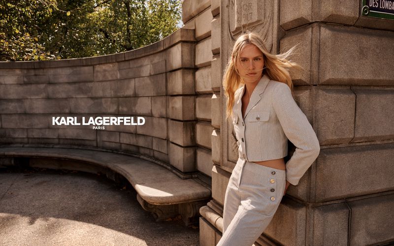 Fashion shot of a blond woman posing in a pinstripe suit in front of a stone wall and leafy green trees in an outdoor setting.
