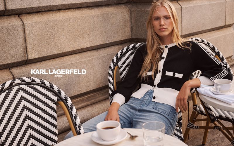 Elegant blonde woman in a black and white cardigan sitting outdoors at a cafe, holding a cup of coffee, showcasing fashion editorial content.